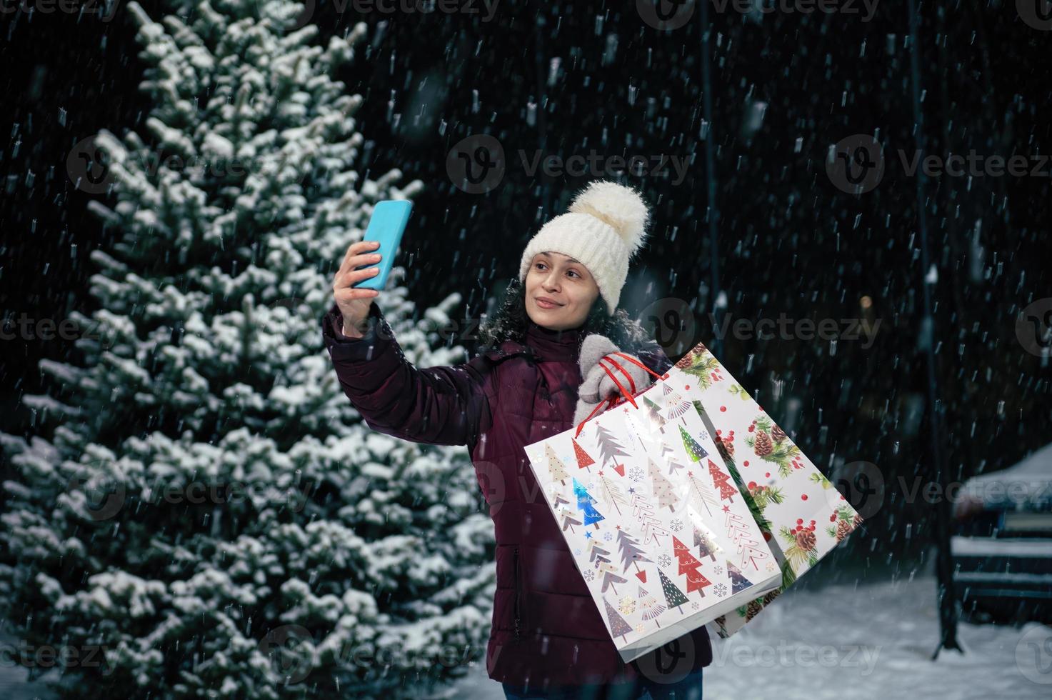 linda mujer toma un selfie en teléfono inteligente, en pie con compras pantalones en contra un cubierto de nieve abeto árbol en un Nevado noche foto