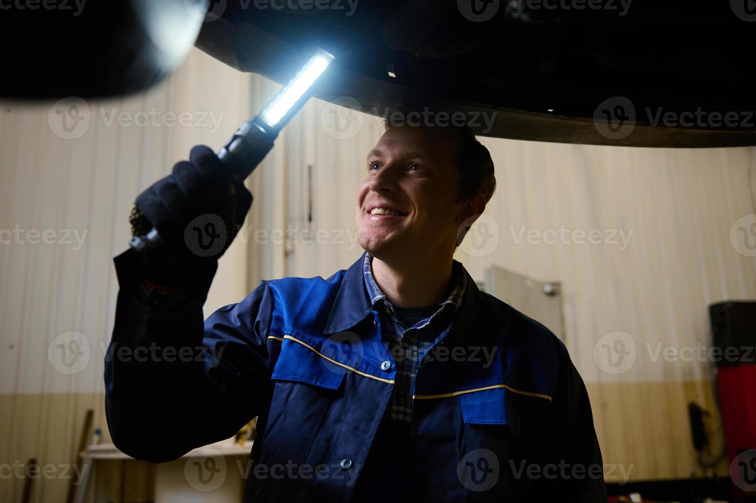 mecánico, técnico, auto ingeniero cheques el debajo de la carrocería de un levantado coche con un lámpara en el garaje de un reparar tienda. coche garantía reparar y mantenimiento, coche Servicio concepto foto