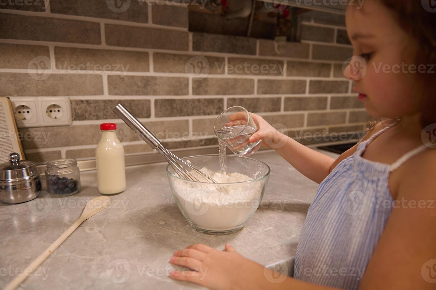 atención en el mano de adorable pequeño niño chica, participación un vaso con agua y torrencial eso dentro un cuenco con harina para preparando líquido masa para carnaval panqueques a hogar cocina. foto