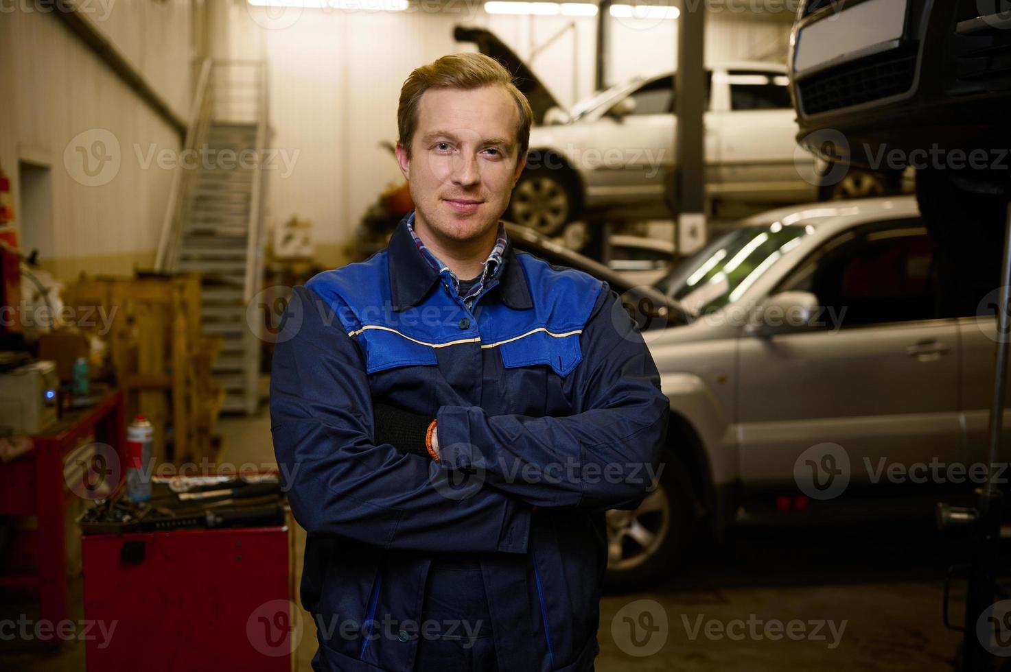 Confident portrait of an auto mechanic dressed in professional uniform looking at camera standing with crossed arms in car repair shop. Car repair, diagnostics and professional service in car service photo