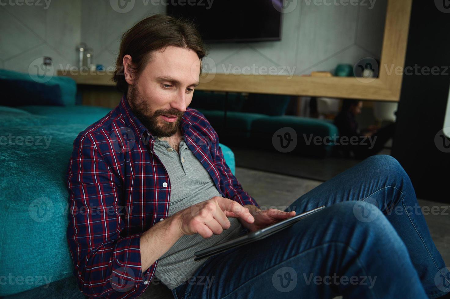 Relaxed smiling Caucasian man in casual denim and checkered shirt reads news on digital tablet, surfs internet, sitting on floor at home leaning back on a sofa. Leisure time, recreation, lifestyle photo