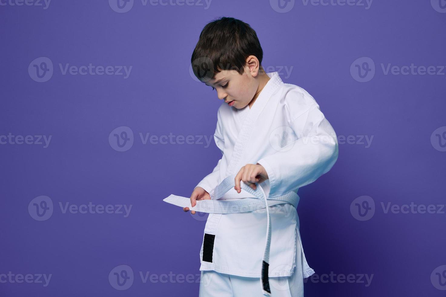 Confident European teenage boy puts on white kimono, ties a belt around his waist, ready for combat sports. Aikido fighter. Oriental martial arts photo