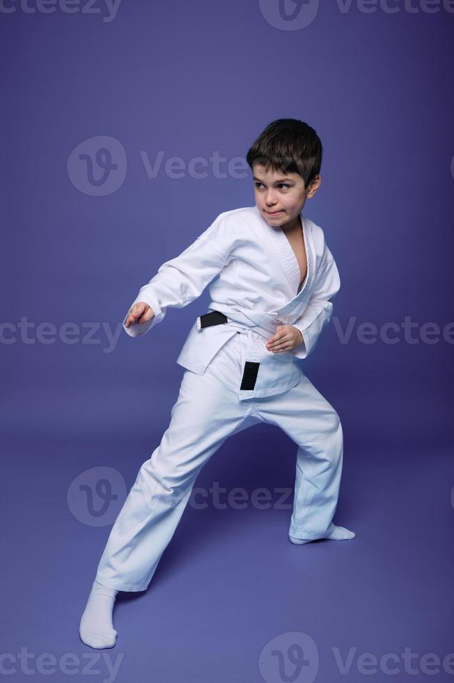 Oriental martial arts. Aikido fighter, Caucasian 10 years old boy in kimono improves his fighting skills, isolated on violet background with copy space photo