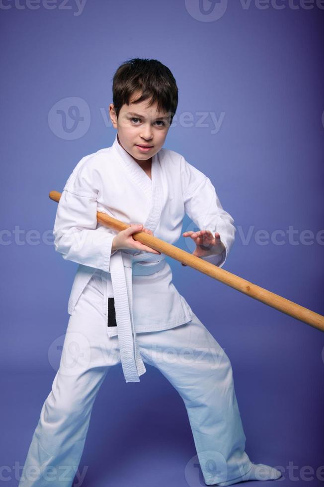 A teenage boy in a white kimono fights with a wooden sword in aikido training on a purple background. Healthy lifestyle and martial arts concept photo