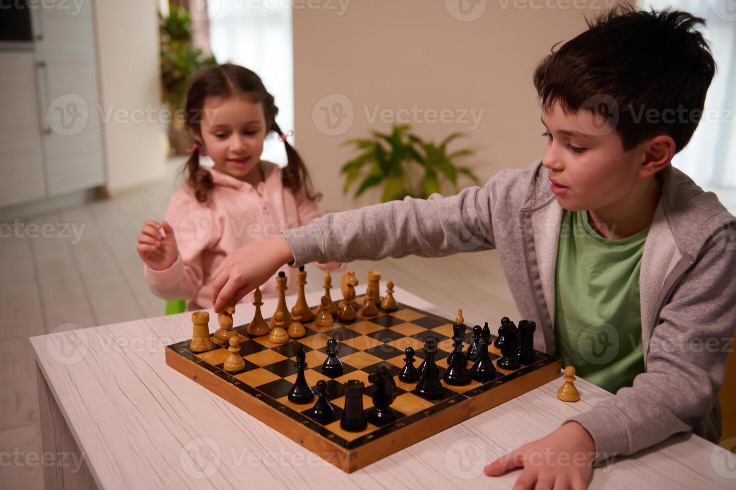 A cute boy with a serious look sits at the table and plays chess with his younger sister, picking up a piece of a knight and making his move. Early development, home educational games for children photo