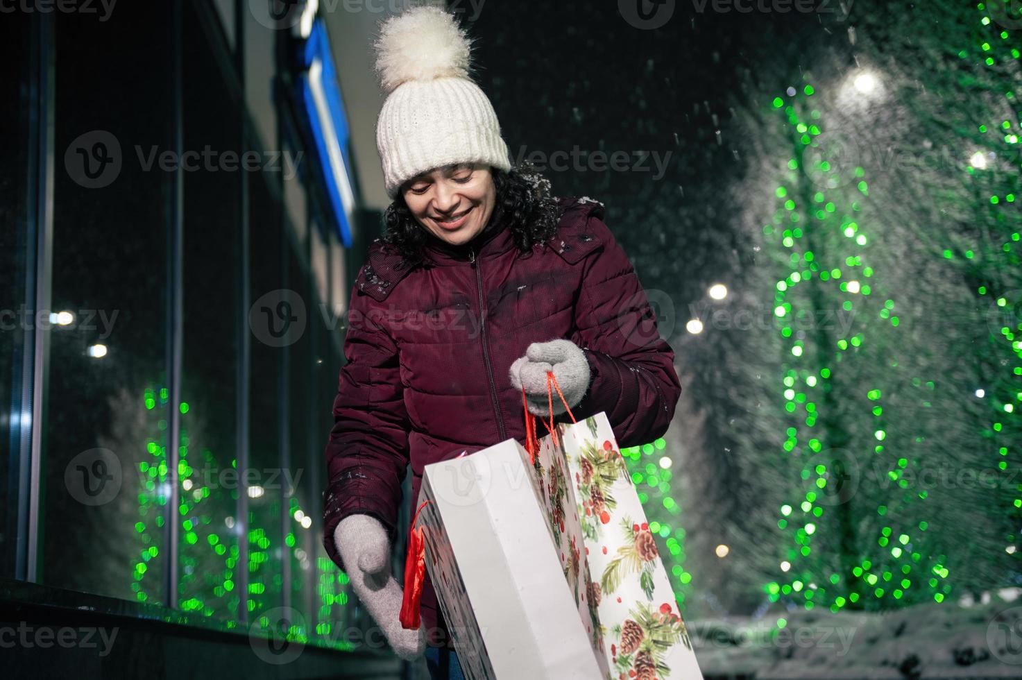 encantador mujer con compras bolsas, caminando abajo el calle, aligerado por festivo iluminación a invierno Nevado noche. foto