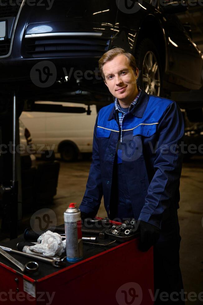 profesional retrato de un hermoso caucásico hombre, auto mecánico, técnico, coche ingeniero en pie cerca un caja con conjunto de herramientas para reparando un levantado automóvil en un izar en el reparar tienda garaje foto