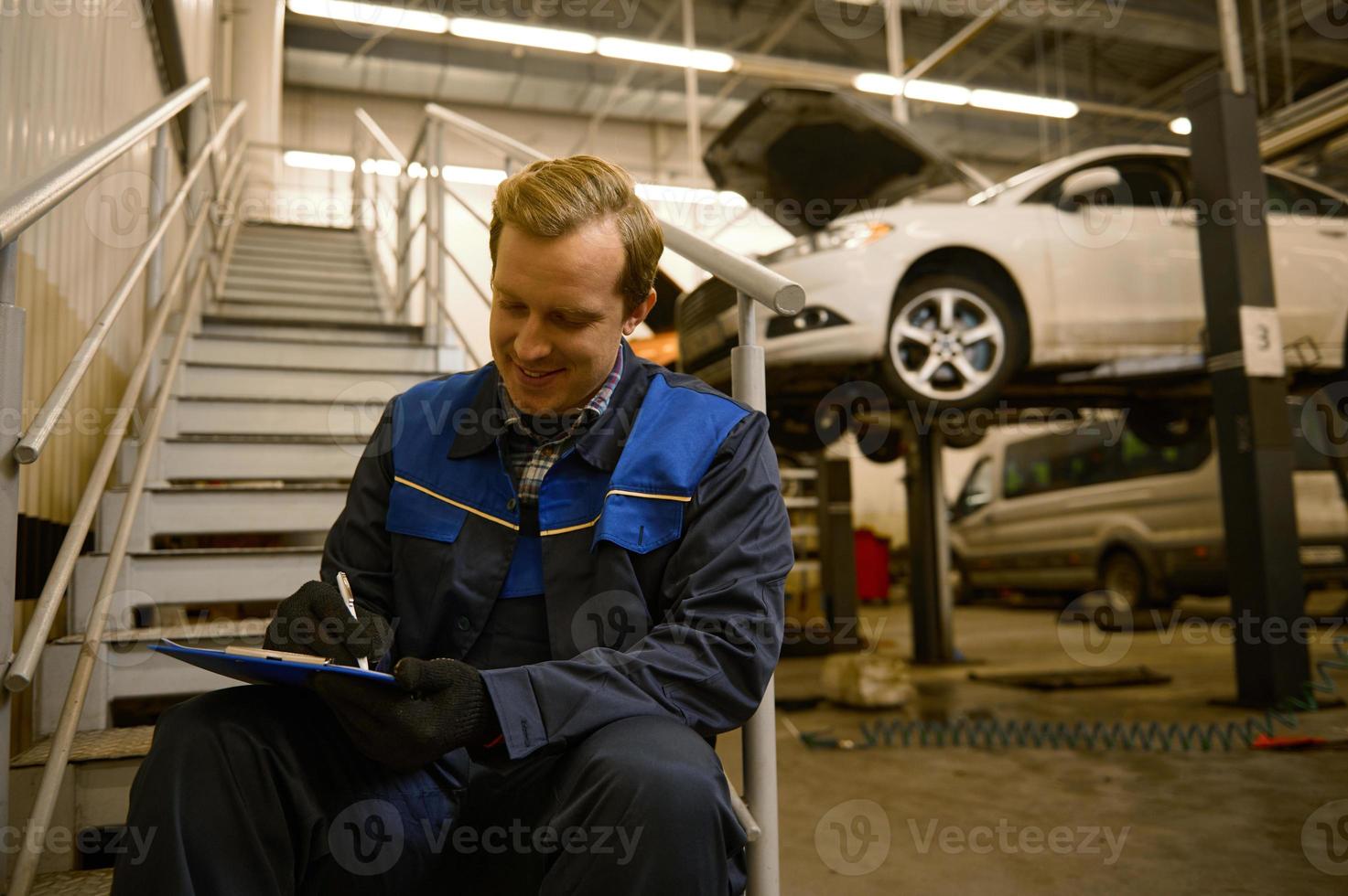 joven caucásico auto mecánico sentado en escalera en el reparar tienda y escritura listas de verificación en portapapeles, inspeccionando el coche en taller. mecánico mantenimiento ingeniero trabajando en automotor industria. foto