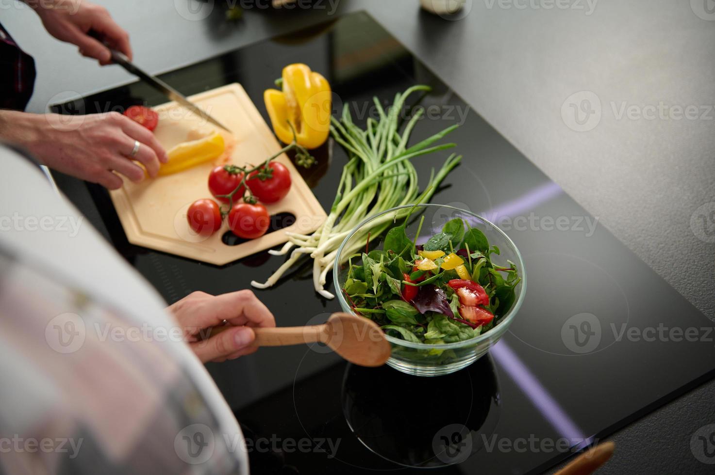 Close-up of a glass bowl with fresh raw salad and male hands slicing yellow bell pepper and tomatoes on a cutting board and pregnant woman's hands holding wooden spoon for mixing ingredients photo