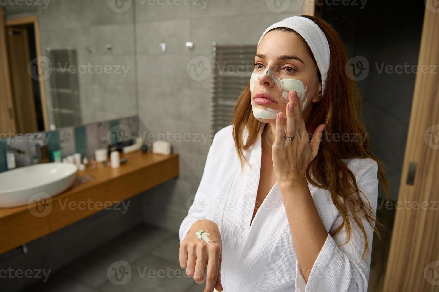 atractivo embarazada rojo peludo mujer en blanco bata de baño en pie en el baño y tomando cuidado de su cara piel, aplicando hidratante o facial exfoliante fregar, haciendo suavizado masaje movimientos foto