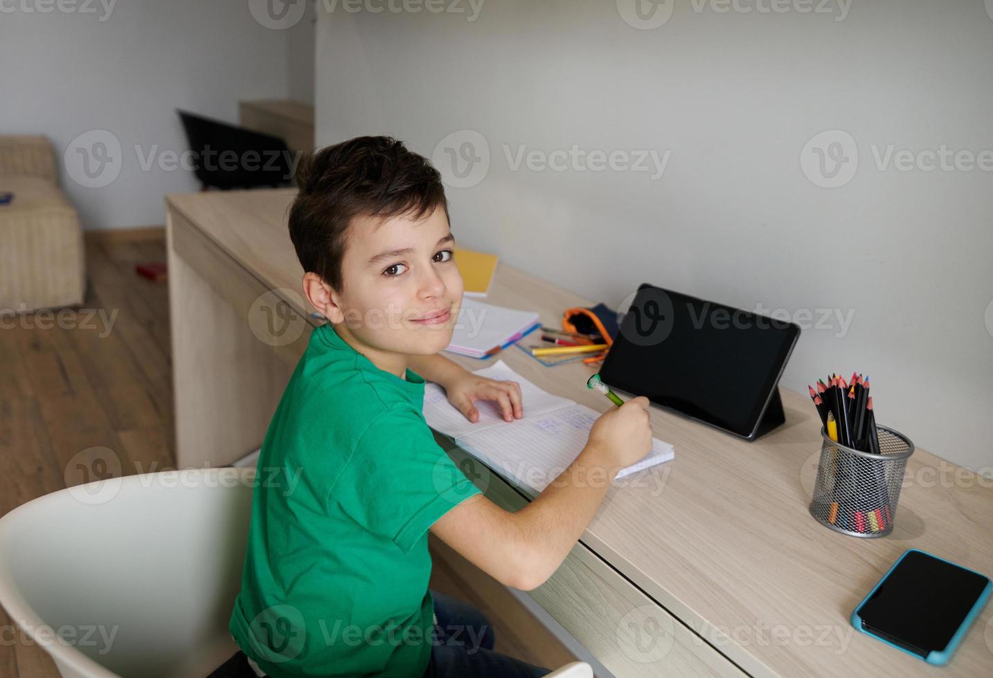 Handsome Caucasian 9 years old school boy sits at wooden desk in front of a digital gadget and doing his homework, cutely smiles looking at camera. Education and school concept. Online learning photo