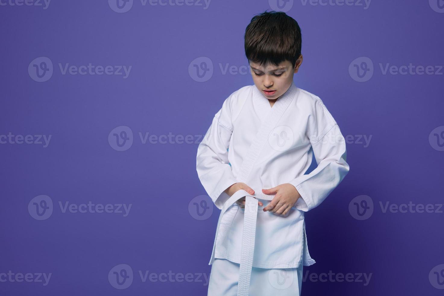 Horizontal portrait of a confident teenage boy aikido wrestler tying white belt of his kimono, isolated over violet background with copy ad space photo