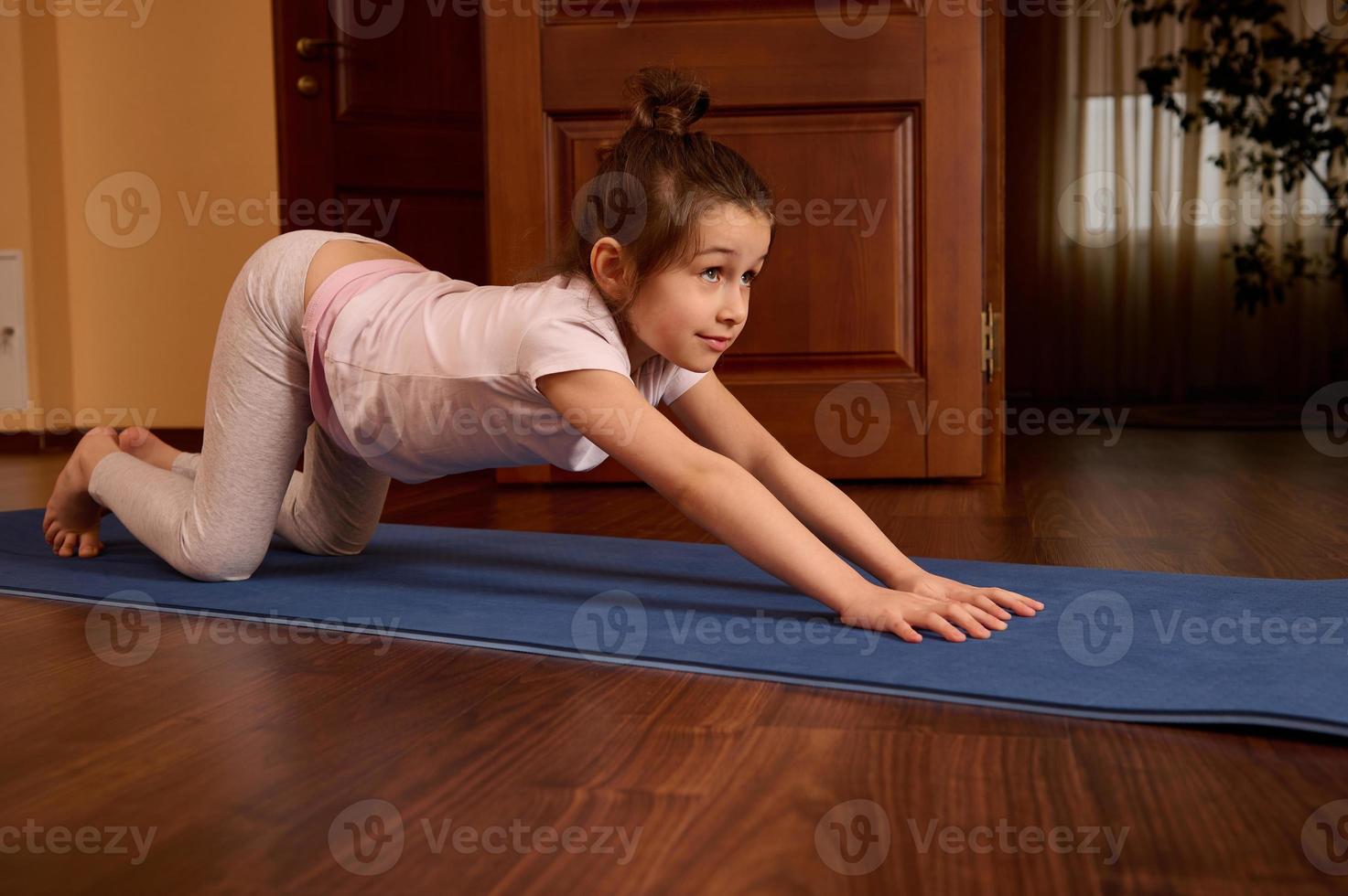 Side portrait of active sporty child girl stretching body and back on fitness mat, practicing yoga, exercising indoors photo