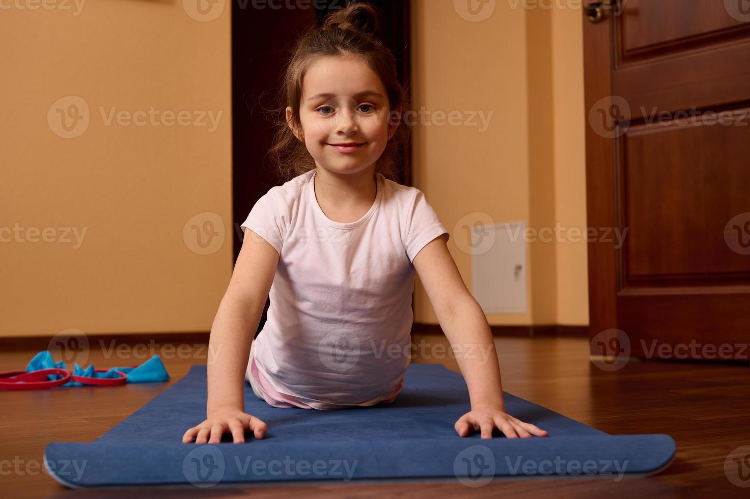 Beautiful little kid girl smiling looking at camera, stretching in upward facing dog pose while practicing yoga on a mat photo