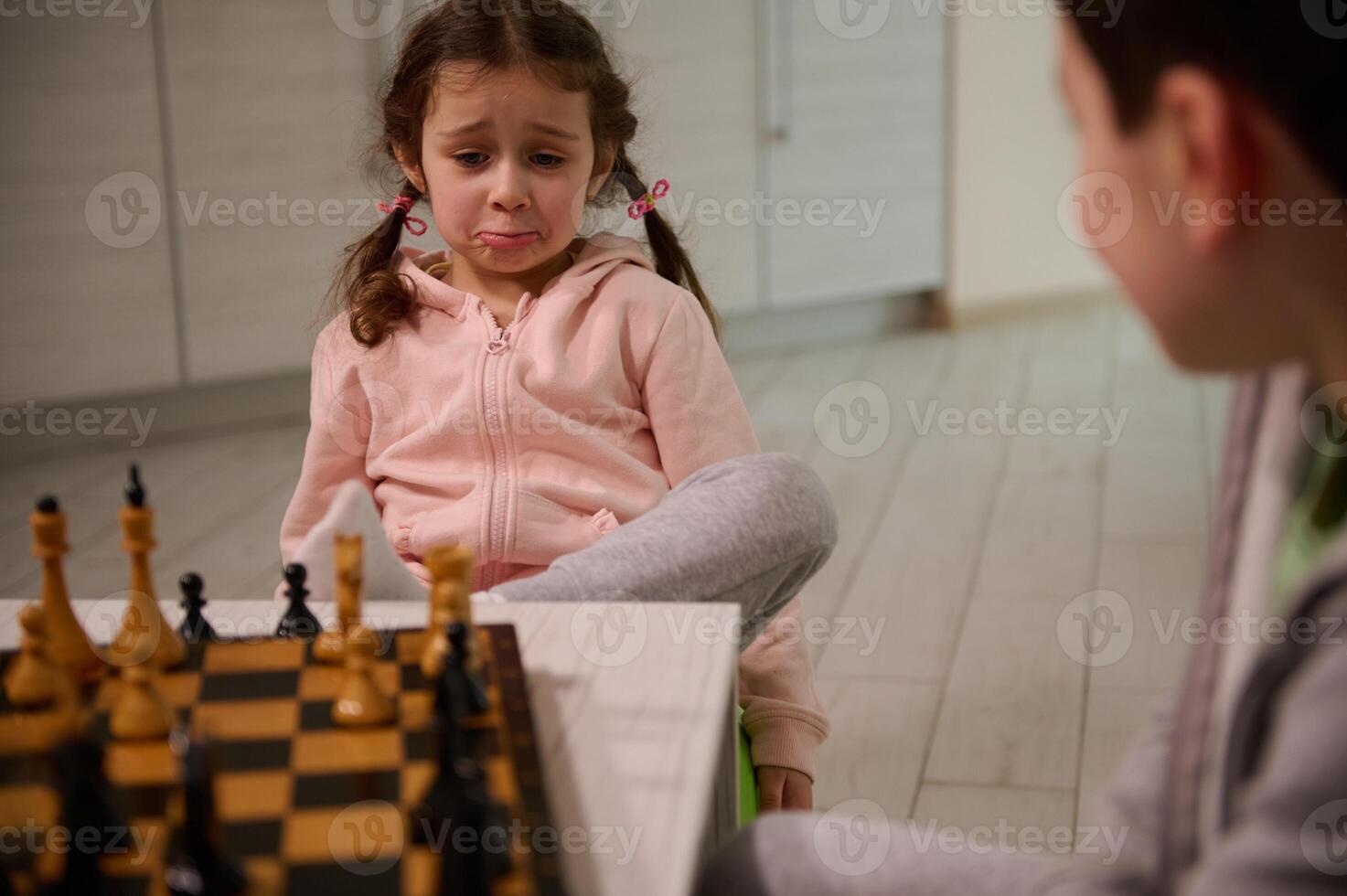 Pretty 4 years old little girl with upset look sitting next to her brother and learning chess game photo