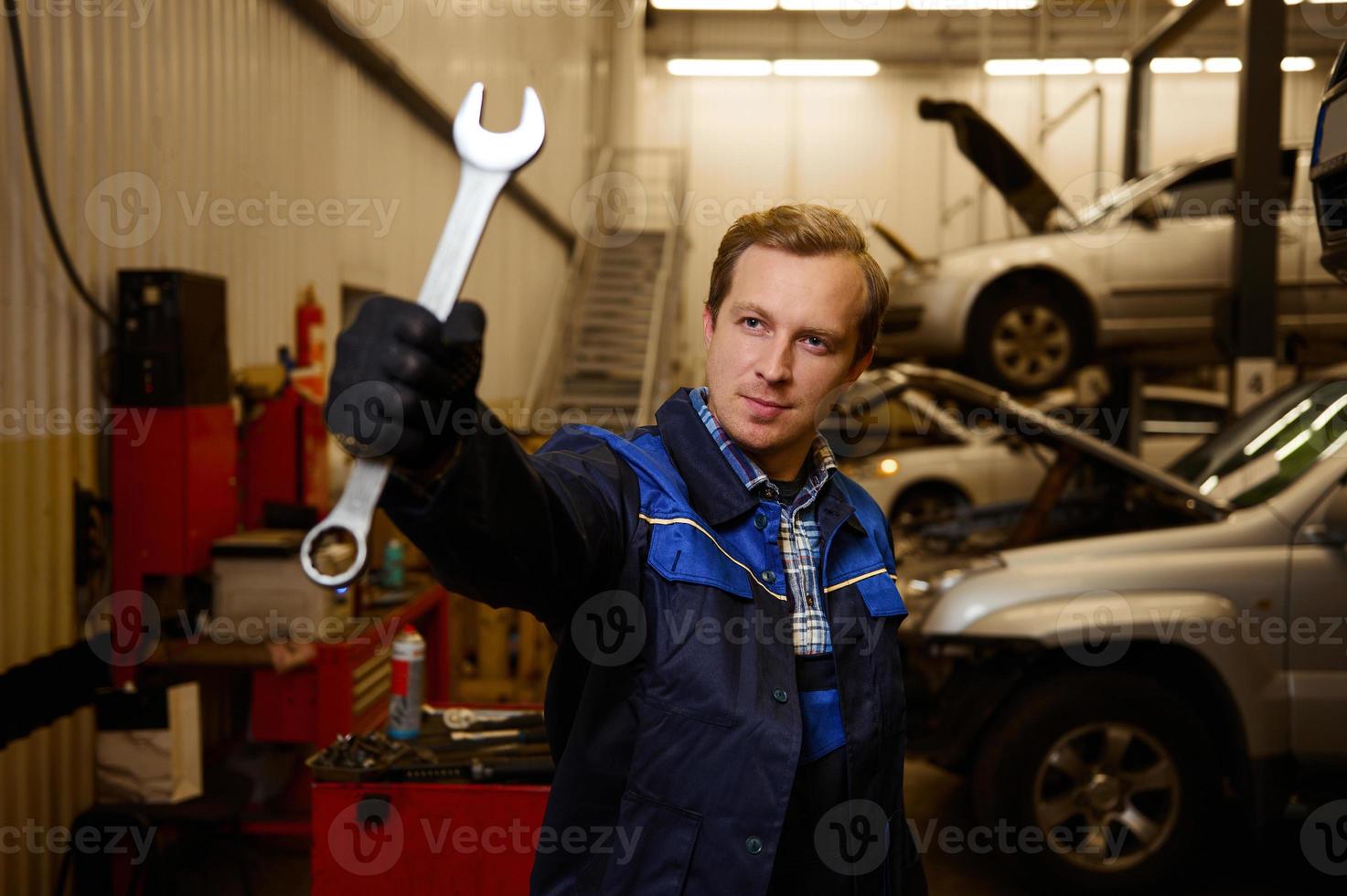 profesional retrato de un joven caucásico auto mecánico en uniforme participación un llave inglesa mientras en pie a su lugar de trabajo en un coche servicio. coche reparar y mantenimiento. foto