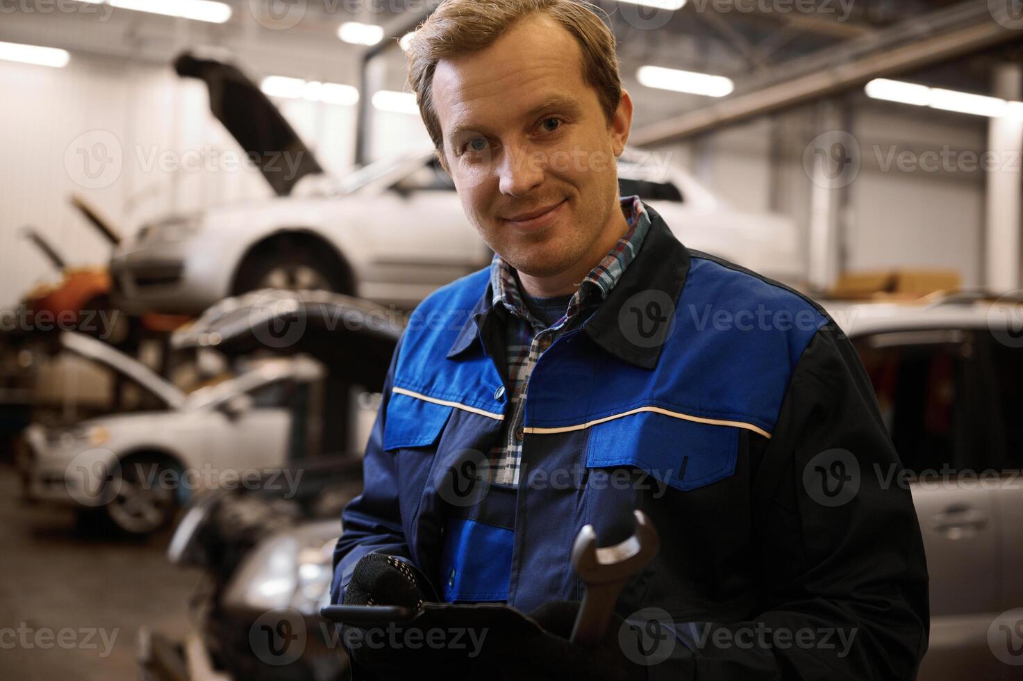 hermoso sonriente caucásico auto mecánico, técnico en uniforme, participación un llave inglesa y en pie en contra levantado carros con abierto capucha en el reparar tienda foto