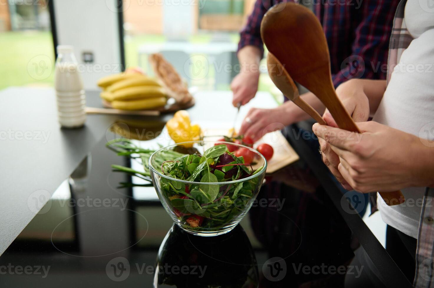 Focus on a glass bowl full of healthy greens, lettuce leaves and fresh vegetables on the background of a happy pregnant couple preparing healthy raw vegan salad on the kitchen island photo