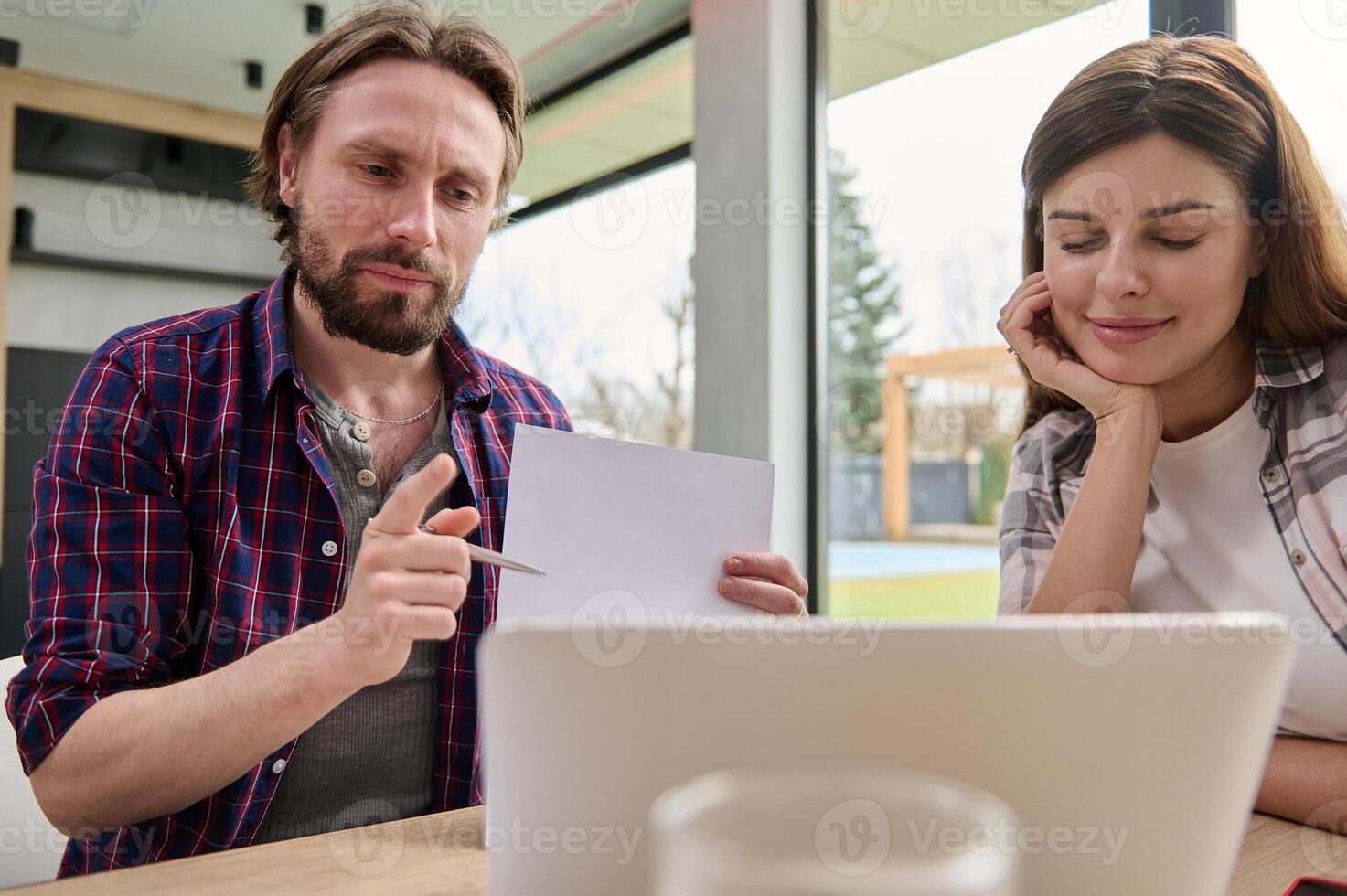 Handsome Caucasian man pointing with a pen on a copy space on the white blank paper sheet while talking by video call sitting at the kitchen table in front of his laptop next to his wife at home photo