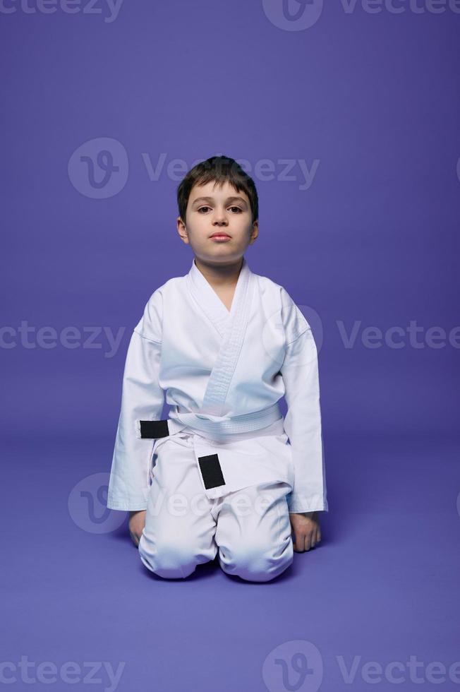 Portrait of a handsome child boy wearing kimono and sitting in aikido stance against purple background with copy space for text. Oriental martial arts concept photo