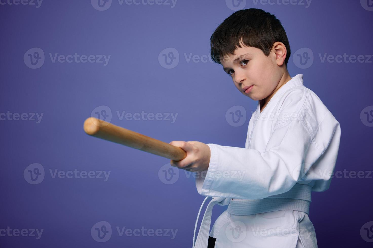 Waist length portrait of a confident teenage boy in white kimono practices wooden bokken in Aikido training on purple background with copy ad space photo