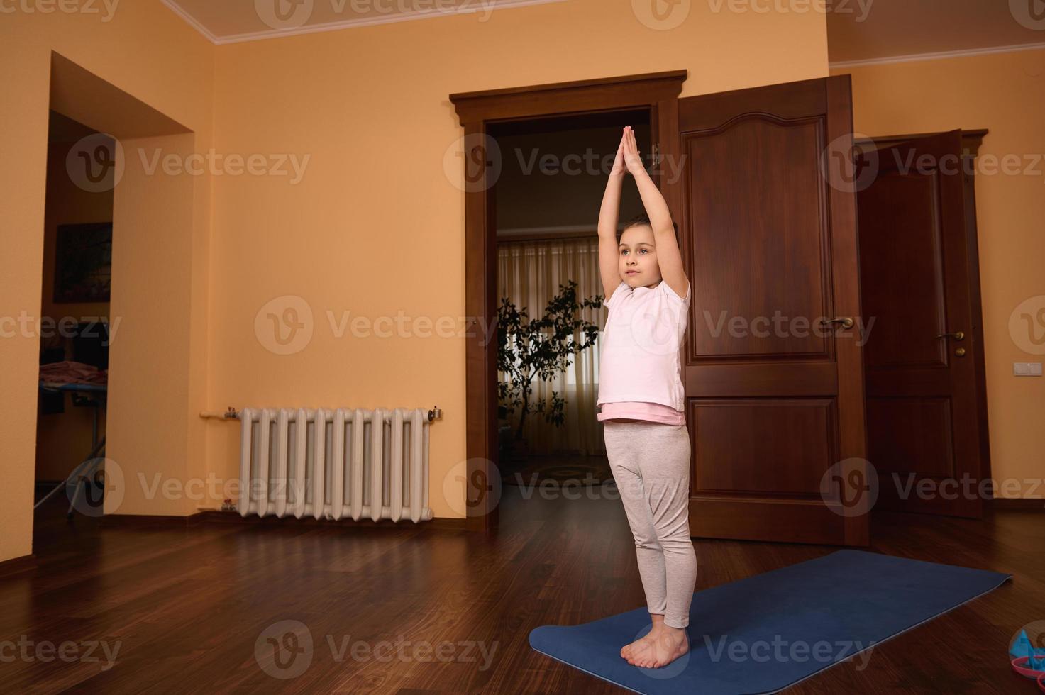 Lovely little kid girl exercising on yoga mat, raising her arms up while stretching her body at cozy homely atmosphere photo