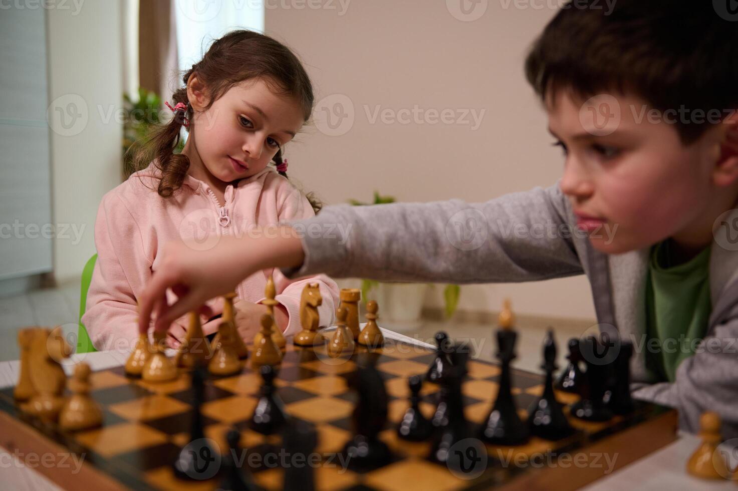 Beautiful little girl looking at her brother moving his chess piece over chessboard, making his move while playing chess game together at home photo
