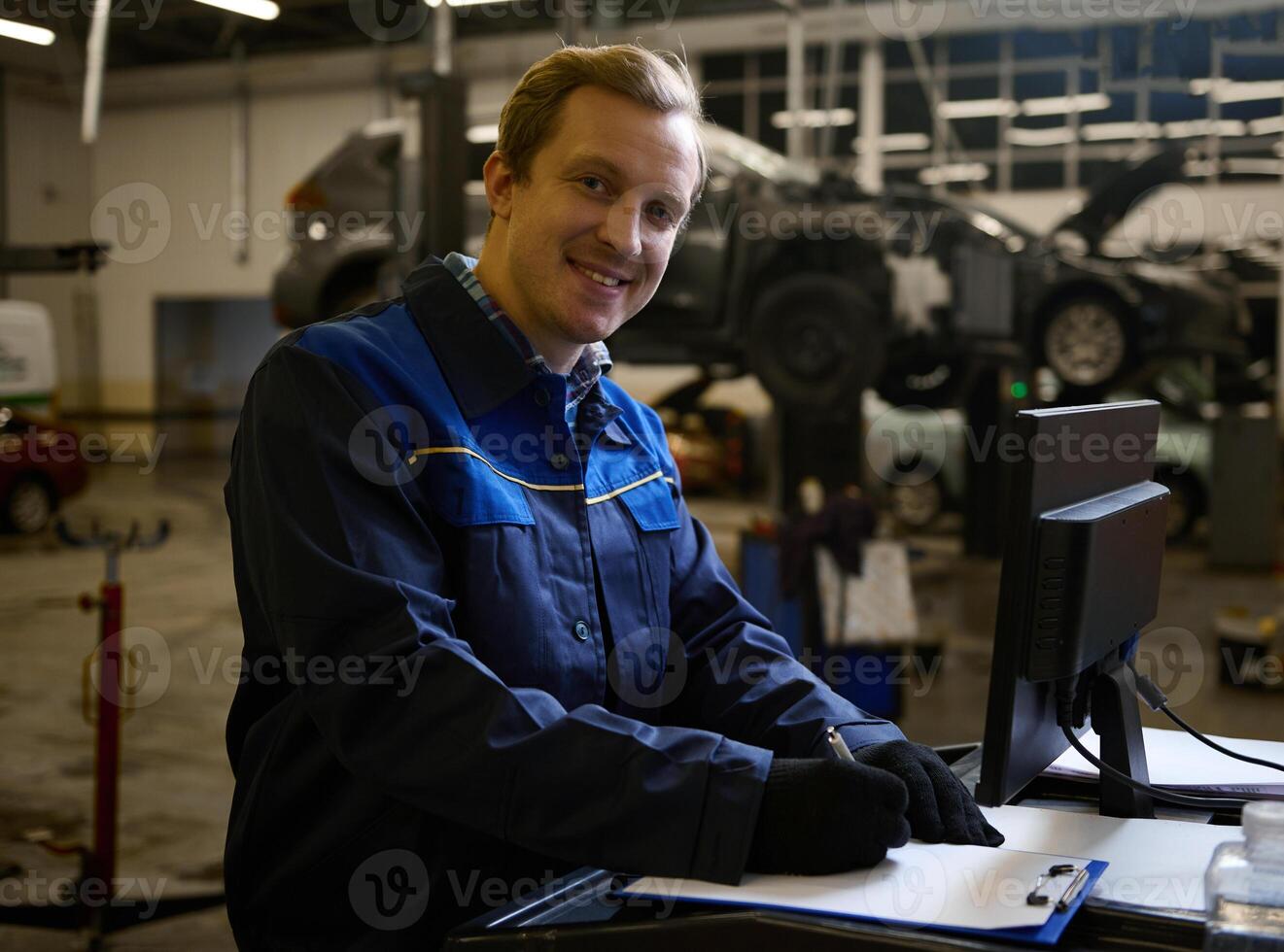Concentrated auto mechanic technician car engineer smiles looking at camera writing checklist on clipboard and working on monitor while servicing a car in the auto service photo