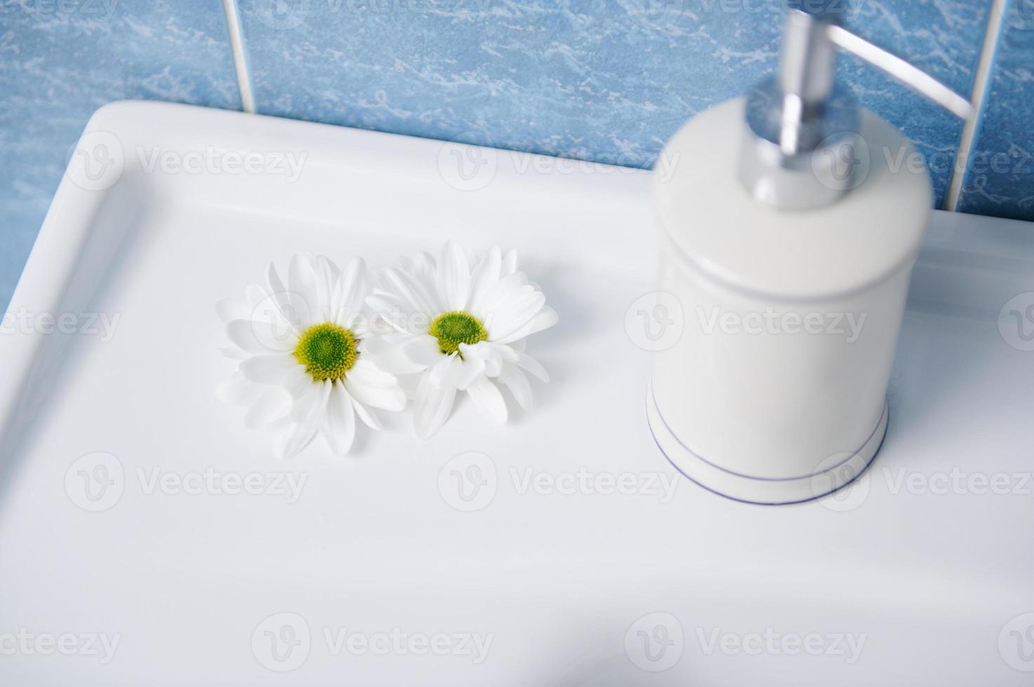 elegante blanco cerámico dispensador con antiséptico antibacteriano líquido jabón y manzanilla flores en un blanco lavar cuenca foto