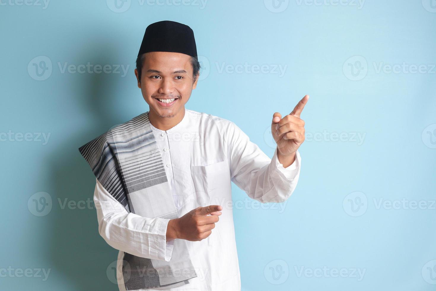 Portrait of young Asian muslim man showing product and pointing with his hand and finger to the side. Isolated image on blue background photo