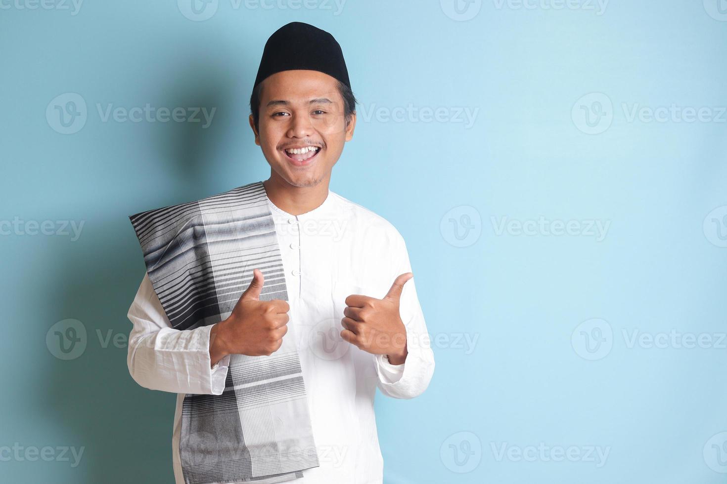 Portrait of Asian muslim man in white koko shirt with skullcap showing thumb up, good job hand gesture. Isolated image on gray background photo