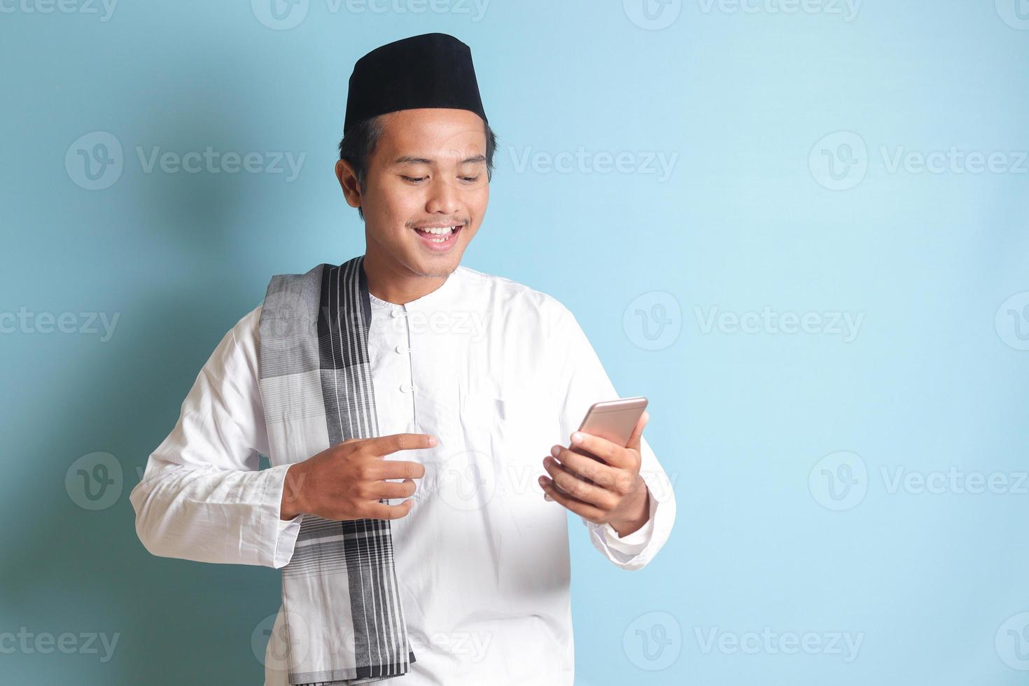 Portrait of young Asian muslim man holding and touching mobile phone with smiling expression on face. Isolated image on blue background photo