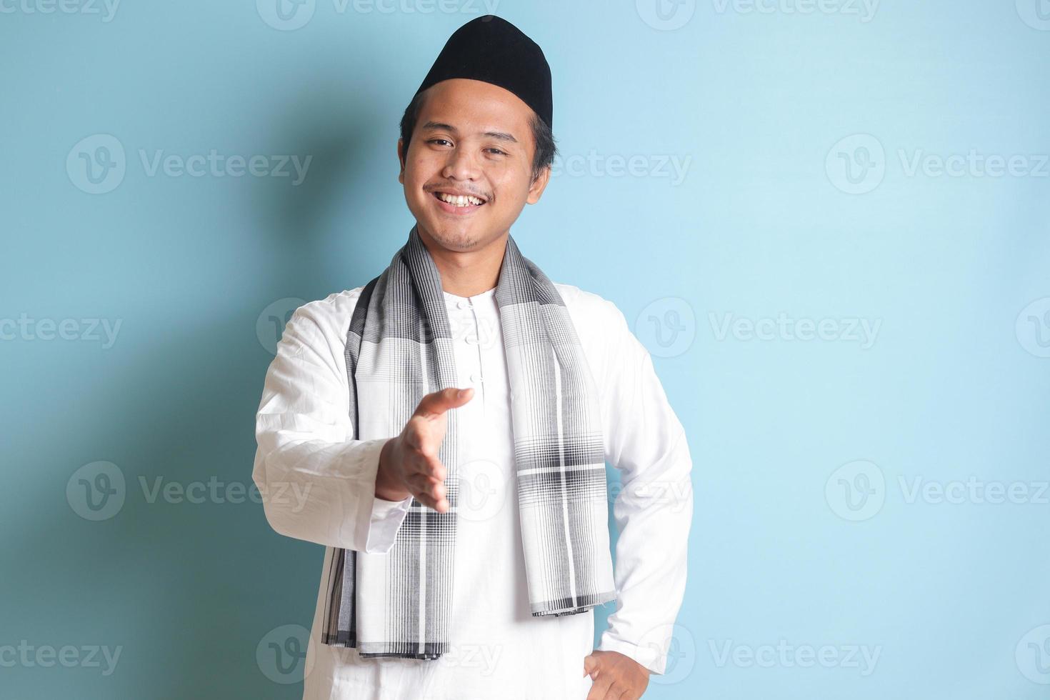 retrato de atractivo asiático musulmán hombre en blanco camisa alcanzando fuera su mano para un apretón de manos, acogedor alguien. aislado imagen en azul antecedentes foto