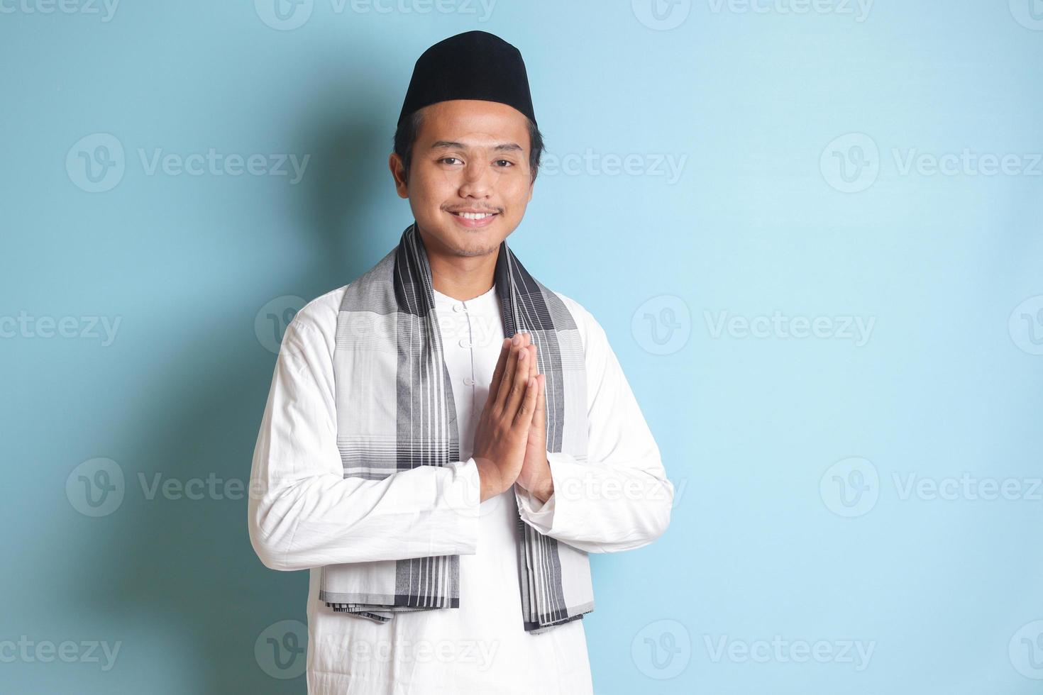 Portrait of Asian muslim man in white koko shirt with skullcap showing apologize and welcome hand gesture. Isolated image on blue background photo