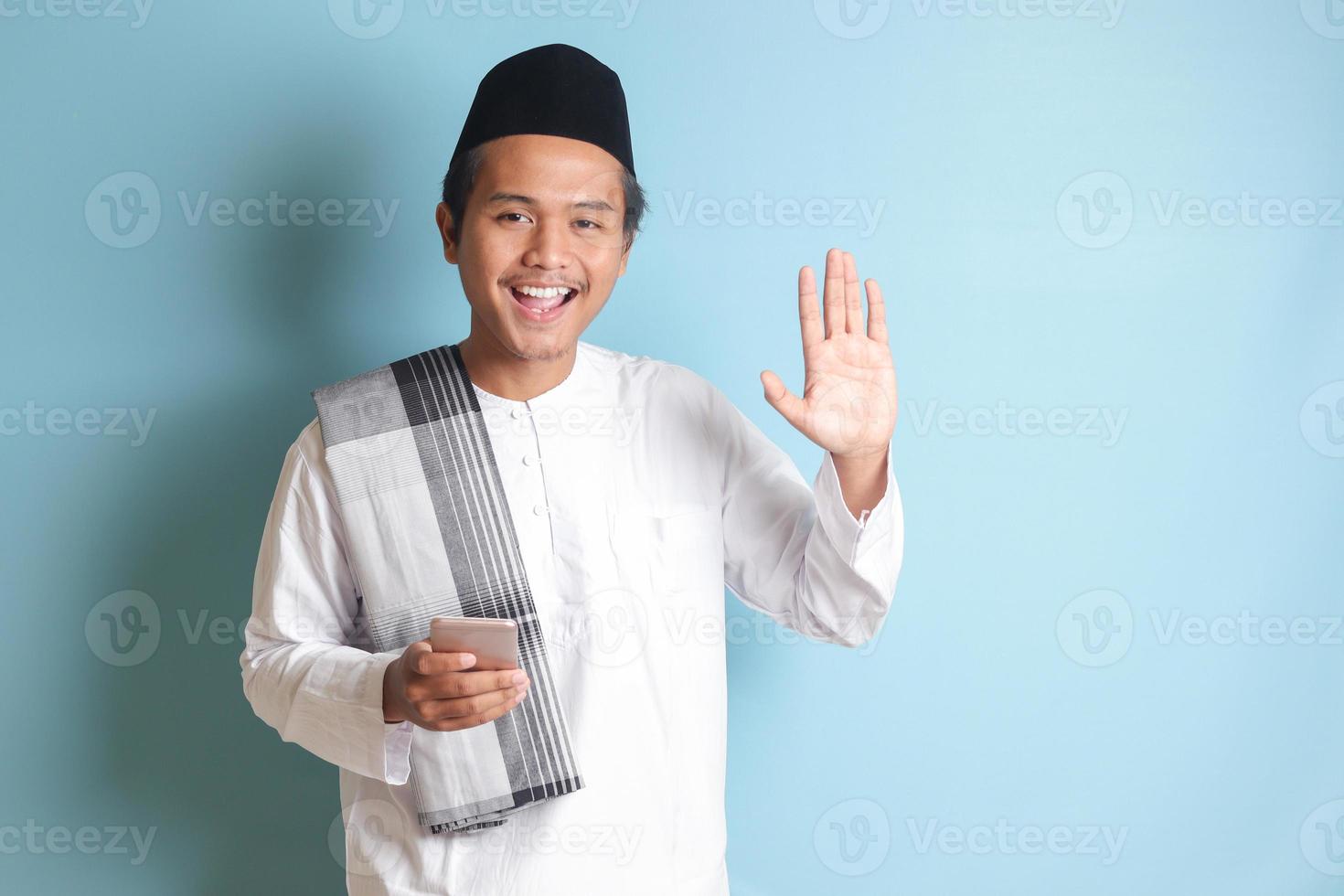 Portrait of young Asian muslim man holding mobile phone with smiling expression on face and waving his hand, saying hi to camera. Isolated image on blue background photo