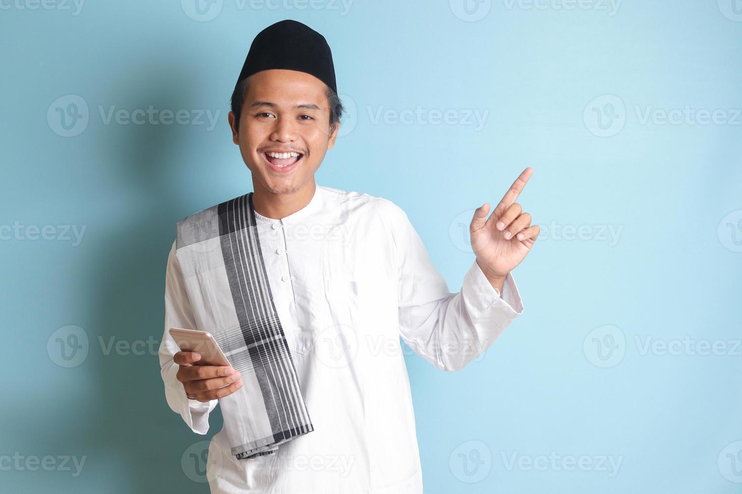 Portrait of young Asian muslim man holding mobile phone with smiling expression on face while pointing finger to the side. Isolated image on blue background photo