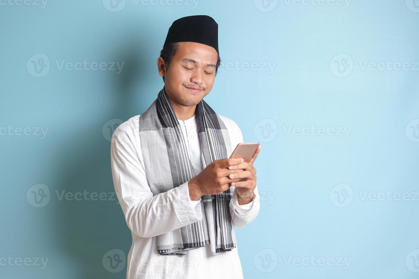 Portrait of young Asian muslim man holding and touching mobile phone with smiling expression on face. Isolated image on blue background photo