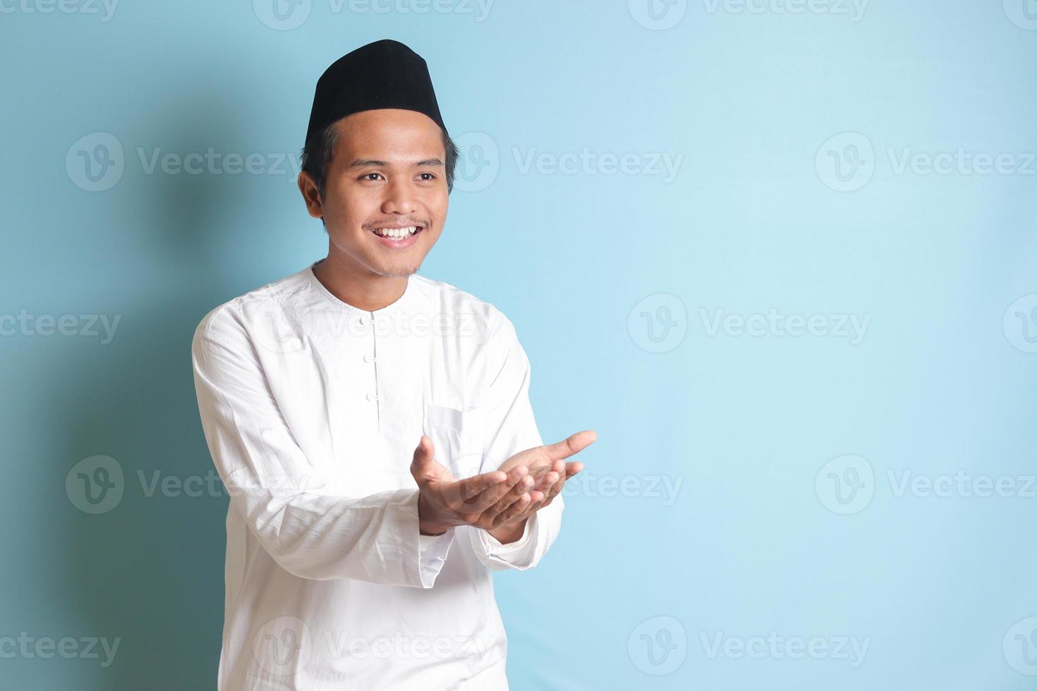 Portrait of Asian muslim man in white koko shirt with skullcap pulling hands towards camera, inviting someone to come inside, welcoming gesture. Isolated image on blue background photo