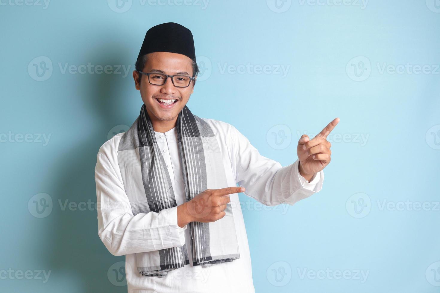 Portrait of young Asian muslim man showing product and pointing with his hand and finger to the side. Isolated image on blue background photo