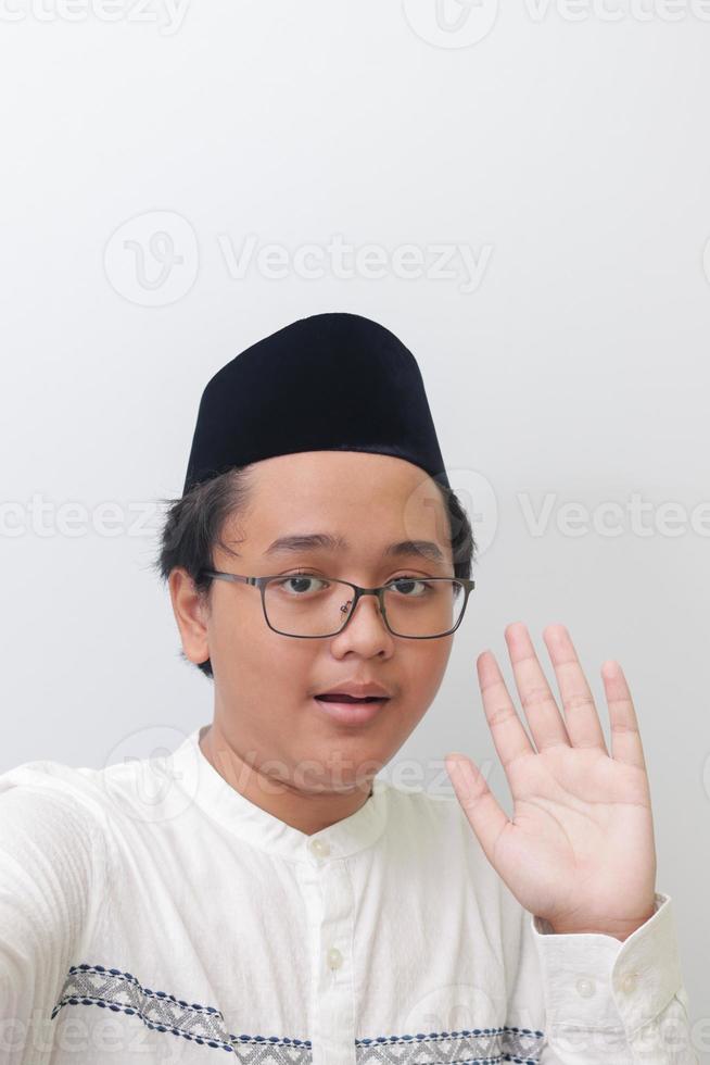 Portrait of smiling young Asian muslim man taking picture of himself or selfie, saying hi and waving his hand. Screen capture of a phone. Isolated image on white background photo