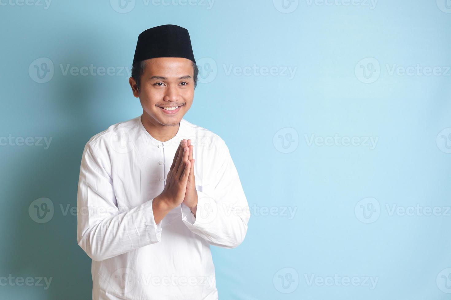 Portrait of Asian muslim man in white koko shirt with skullcap showing apologize and welcome hand gesture. Isolated image on blue background photo