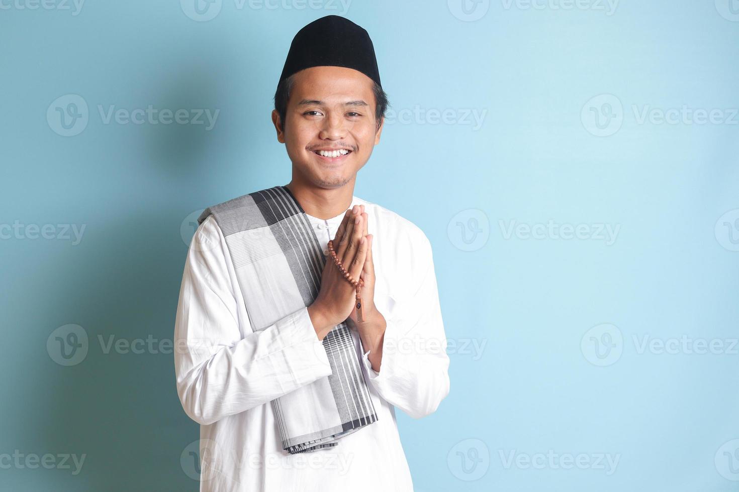 Portrait of Asian muslim man in white koko shirt with skullcap showing apologize and welcome hand gesture. Isolated image on blue background photo