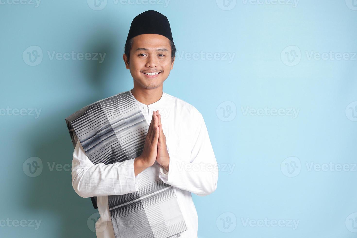 Portrait of Asian muslim man in white koko shirt with skullcap showing apologize and welcome hand gesture. Isolated image on blue background photo