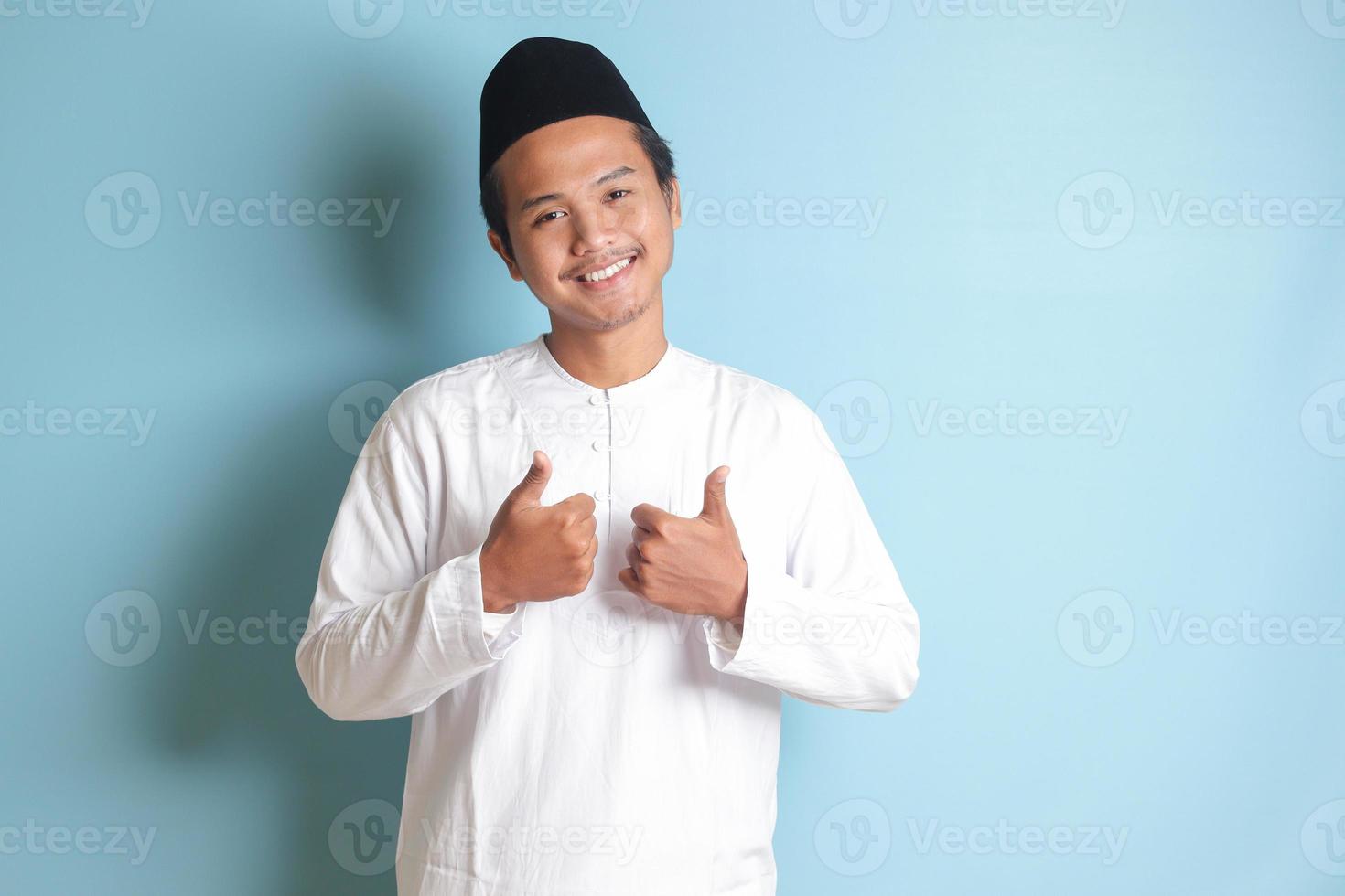 Portrait of Asian muslim man in white koko shirt with skullcap showing thumb up, good job hand gesture. Isolated image on gray background photo