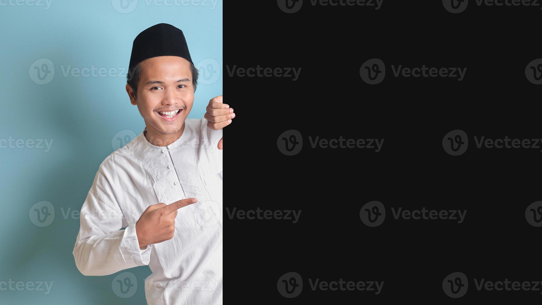 Portrait of young Asian muslim man showing product and pointing finger at copy space on big black paper poster advertising your text. Isolated image on blue background photo