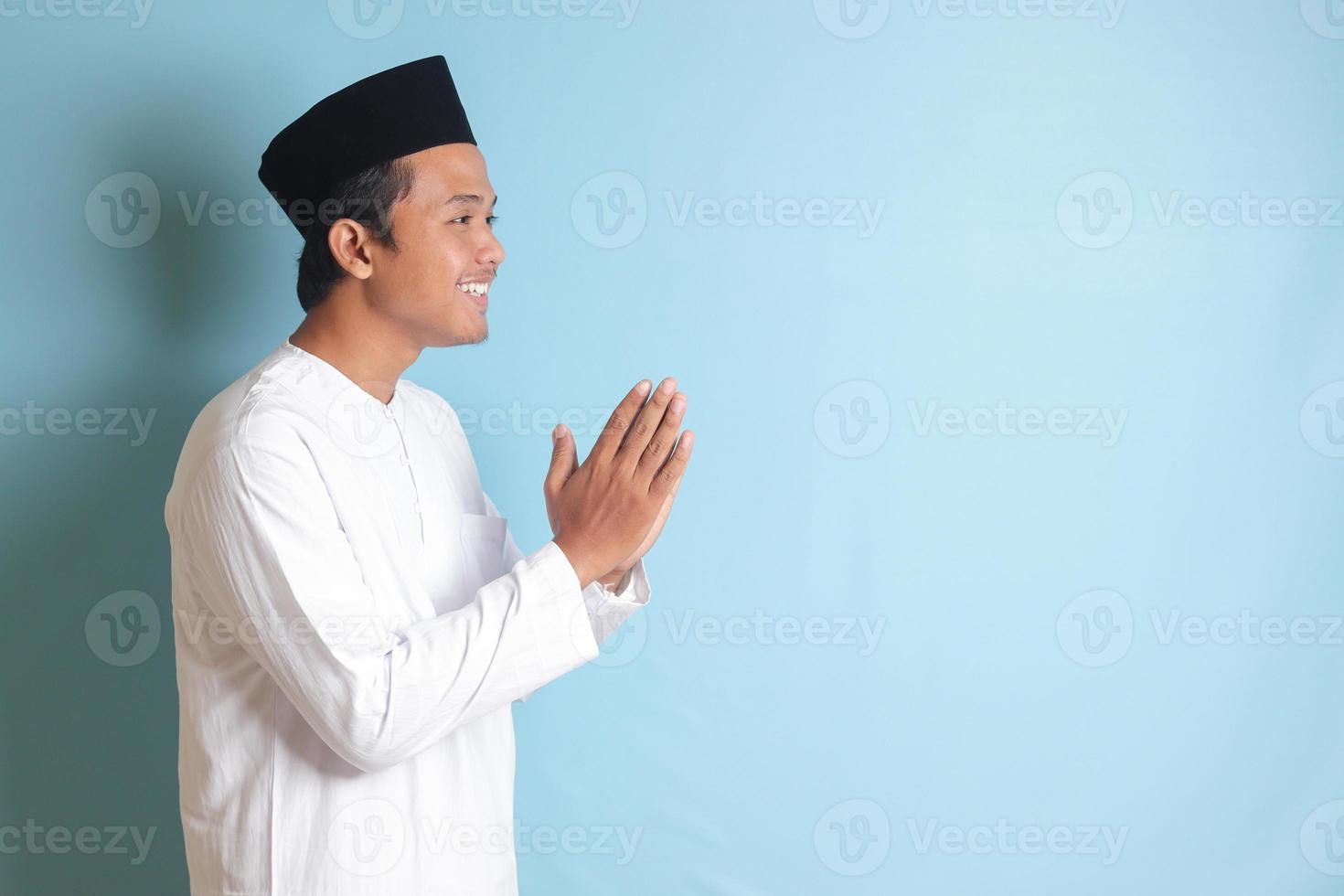 Portrait of Asian muslim man in white koko shirt with skullcap showing apologize and welcome hand gesture. Isolated image on blue background photo