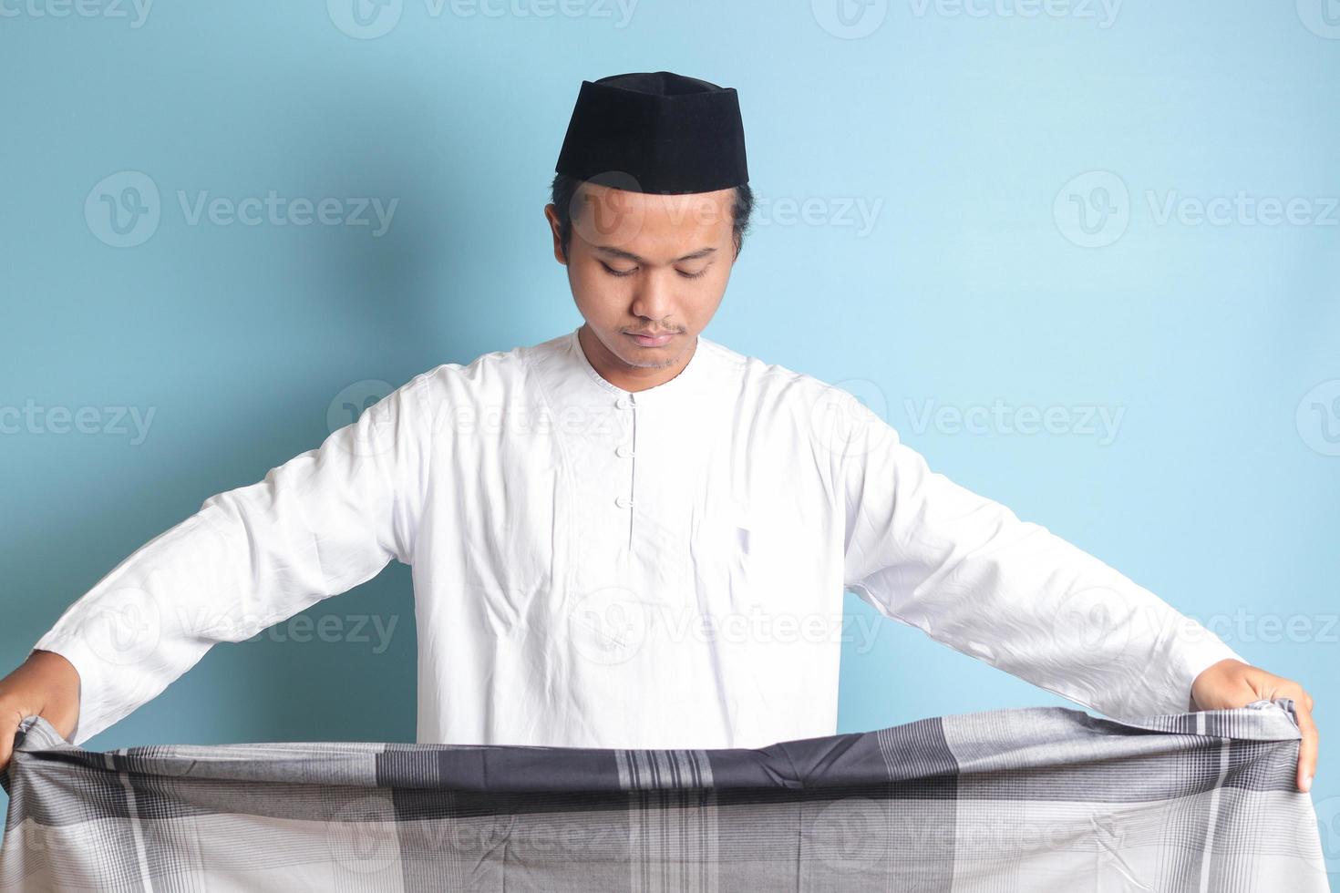 retrato de asiático musulmán hombre en blanco koko camisa plegable y ordenado arriba su Sarung . aislado imagen en azul antecedentes foto