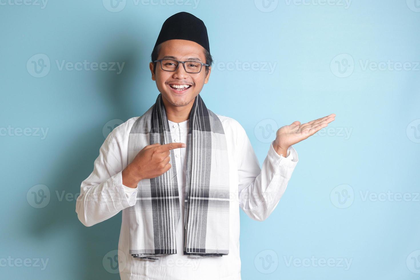 Portrait of young Asian muslim man showing product and pointing with his hand and finger to the side. Isolated image on blue background photo