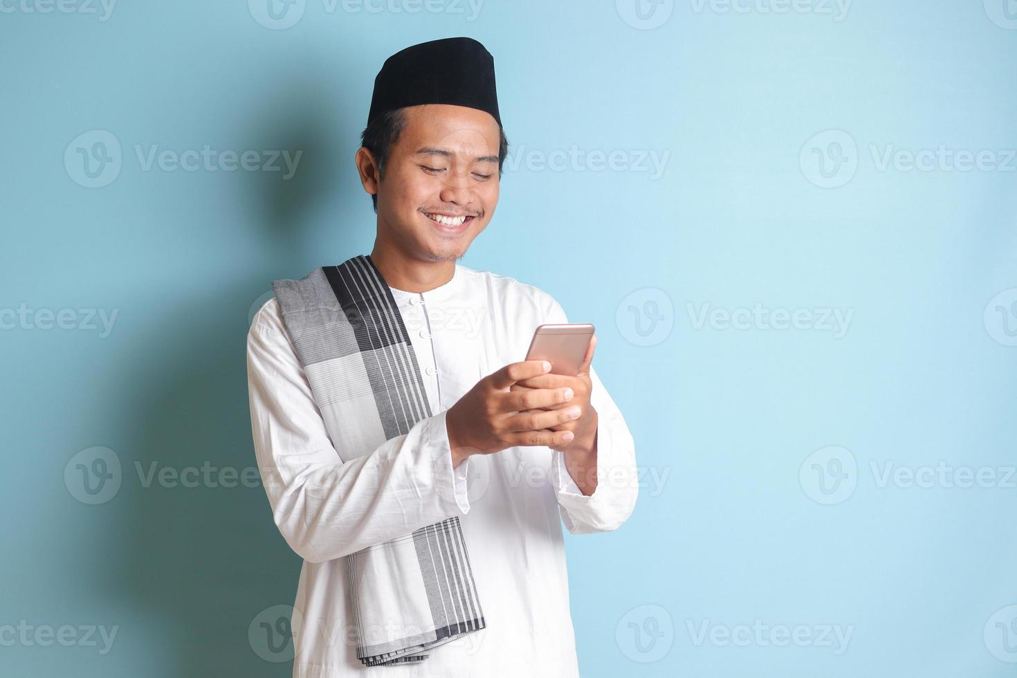 Portrait of young Asian muslim man holding and touching mobile phone with smiling expression on face. Isolated image on blue background photo