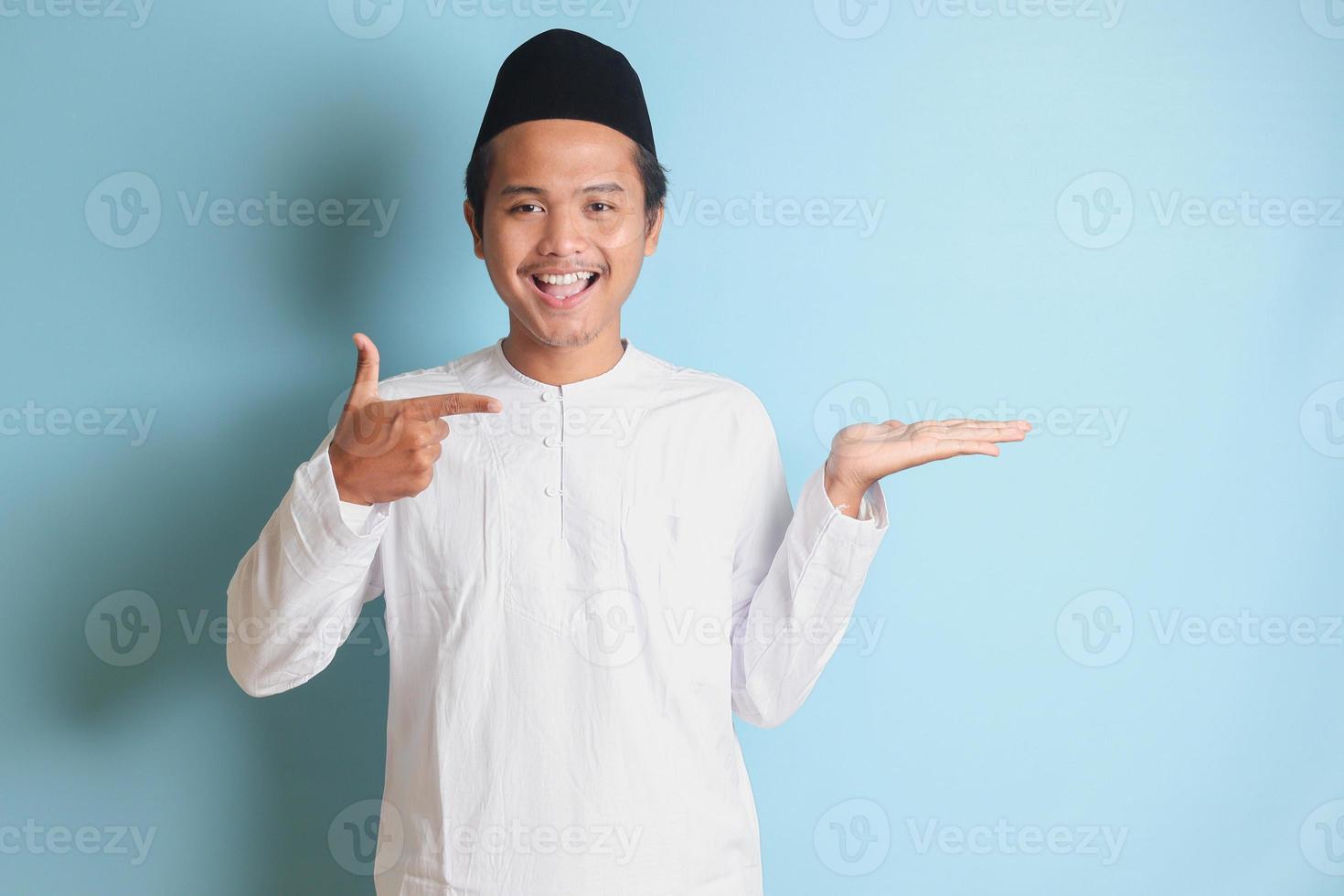 Portrait of young Asian muslim man showing product and pointing with his hand and finger to the side. Isolated image on blue background photo
